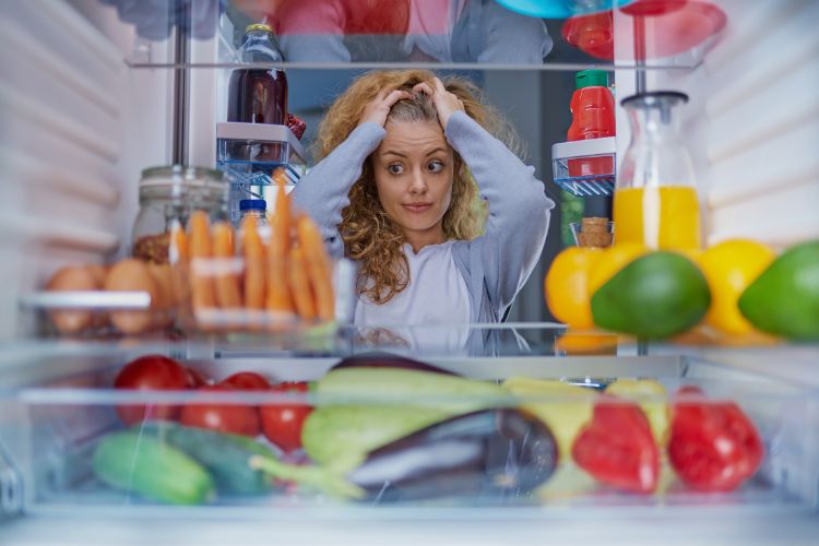 woman standing in the open door of a refrigerator looking at food items with hands on head used on a blog about the worst foods for bloating