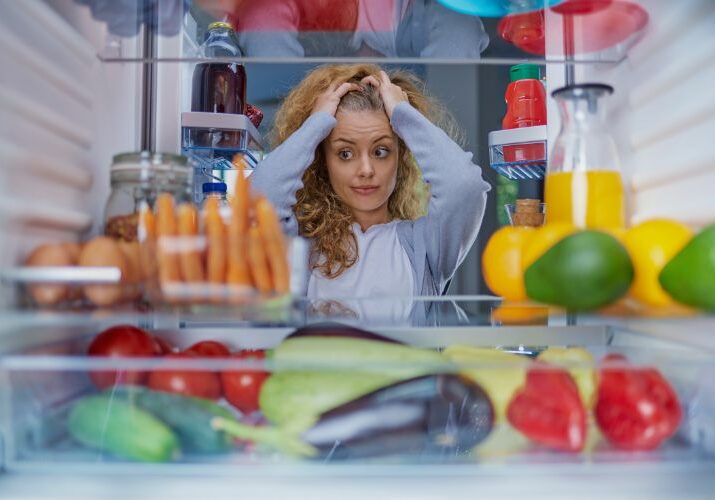 woman standing in the open door of a refrigerator looking at food items with hands on head used on a blog about the worst foods for bloating